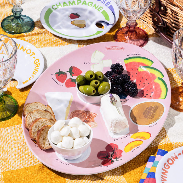 The Summer Soirée Edit by Maple features a pink melamine plate with bread, cheese, olives, blackberries, marshmallows, and watermelon on a yellow plaid cloth, complete with glassware and a picnic basket nearby.