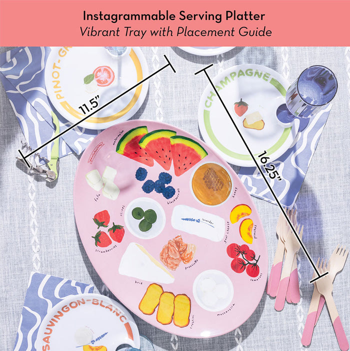 The Maple Meg Quinn Melamine Serving Tray, a pink oval tray with illustrated food pairings and “Pinot Grigio,” “Champagne,” and “Sauvignon Blanc” labels, is dishwasher safe and shown on a table with blue napkins, wooden utensils, and colorful plates.