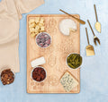 The Maple Meg Quinn Rectangle Charcuterie Board (18x12") displays cheeses, crackers, pecans, pickles, and frozen grapes on a blue surface with gold cheese knives and a beige napkin—ideal for an elegant charcuterie spread.