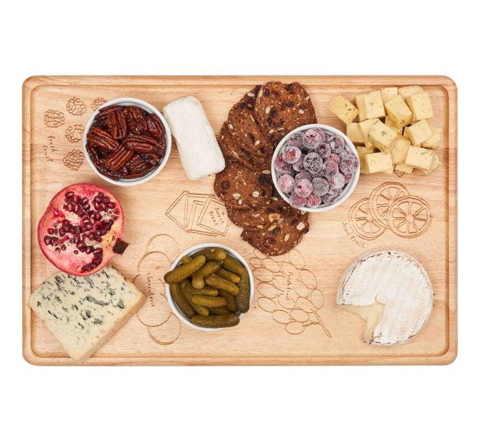The Maple Meg Quinn Rectangle Charcuterie Board (18x12") displays cheeses, crackers, sugared cranberries, candied pecans, cornichons, pomegranate, and bowls on engraved wood sections.