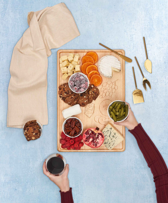 The Maple Meg Quinn Rectangle Charcuterie Board (18x12") holds cheeses, crackers, fruit, pickles, and nuts on a blue table. A hand holds a drink while another reaches for food; gold utensils and a light napkin are beside the board.