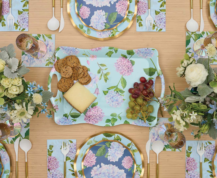 A table set with floral plates, napkins, and placemats, gold utensils, and the Sophistiplate Melamine Hydrangea Serving Tray filled with cheese, crackers, and grapes. Floral centerpieces adorn the light wood surface.