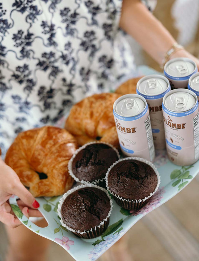 A person holds a Sophistiplate Melamine Hydrangea Serving Tray with two croissants, three chocolate muffins, and four cans of La Colombe draft latte while wearing a patterned outfit.