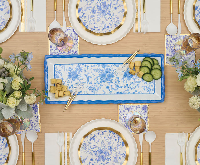 An elegant table setting with blue and white floral plates, gold utensils, clear glasses, a Sophistiplate Melamine Timeless Serving Plate displaying cheese and cucumbers, and floral centerpieces on a light wood surface.