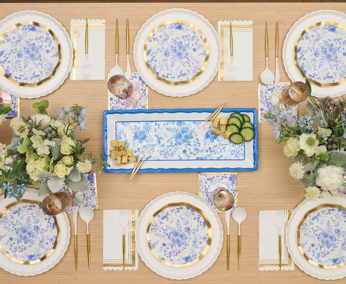 A neatly set table with blue and white floral plates, gold cutlery, floral napkins, and flower centerpieces. At the center, Sophistiplate’s Melamine Timeless Serving Plate holds cheese, cucumber slices, crackers, and gold serving utensils.