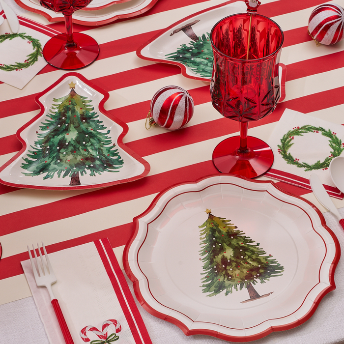 The Merry & Bright: Christmas Table for 16 set includes a red and white striped tablecloth, tree-themed heavy duty plates, red glasses, ornaments, paper napkins, and small wreath illustrations for a cheerful holiday gathering.