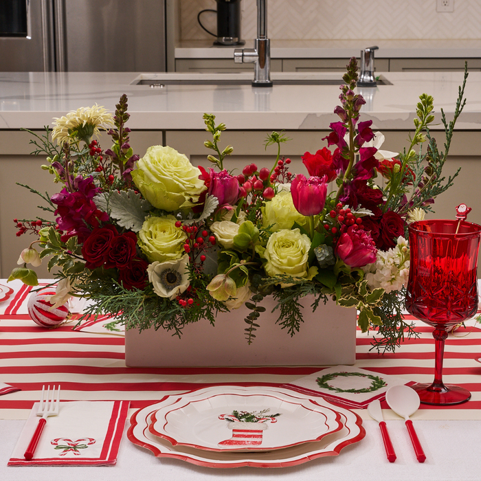 A festive table setting features red and white striped accents, a lush floral centerpiece with roses, tulips, and greenery, vibrant red glassware, and Sophistiplate Merry and Bright Salad Plates (8-pack), along with holiday-themed napkins arranged neatly on the kitchen table.