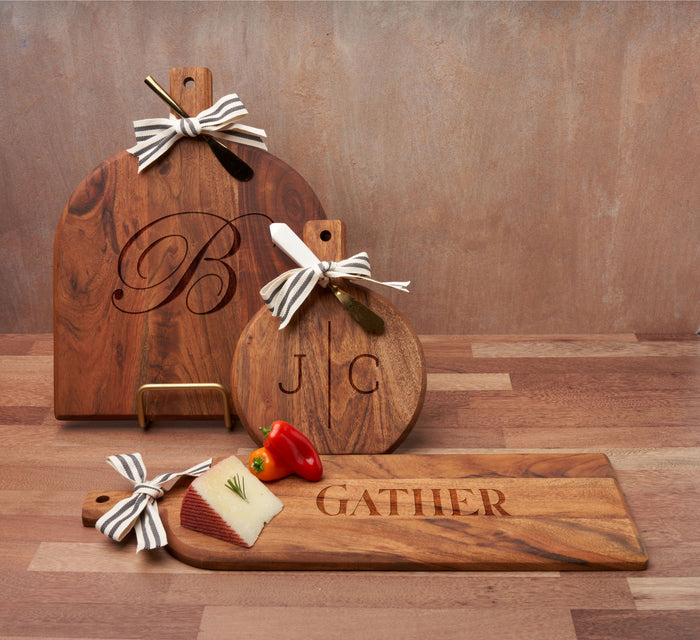 Three wooden cutting boards with bows are shown on a Maple Metal Board Display Stand in matte gold. The boards feature a large B, J | C initials, and "GATHER." Cheese, peppers, and a cheese knife rest on the longest board.