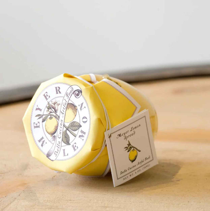 A jar of Bella Cucina Meyer Lemon Spread with a yellow paper cover and a lemon-themed label sits on wood. A tag reading "Meyer Lemon Spread" hangs from it—perfect as a citrusy spread or dessert topping.