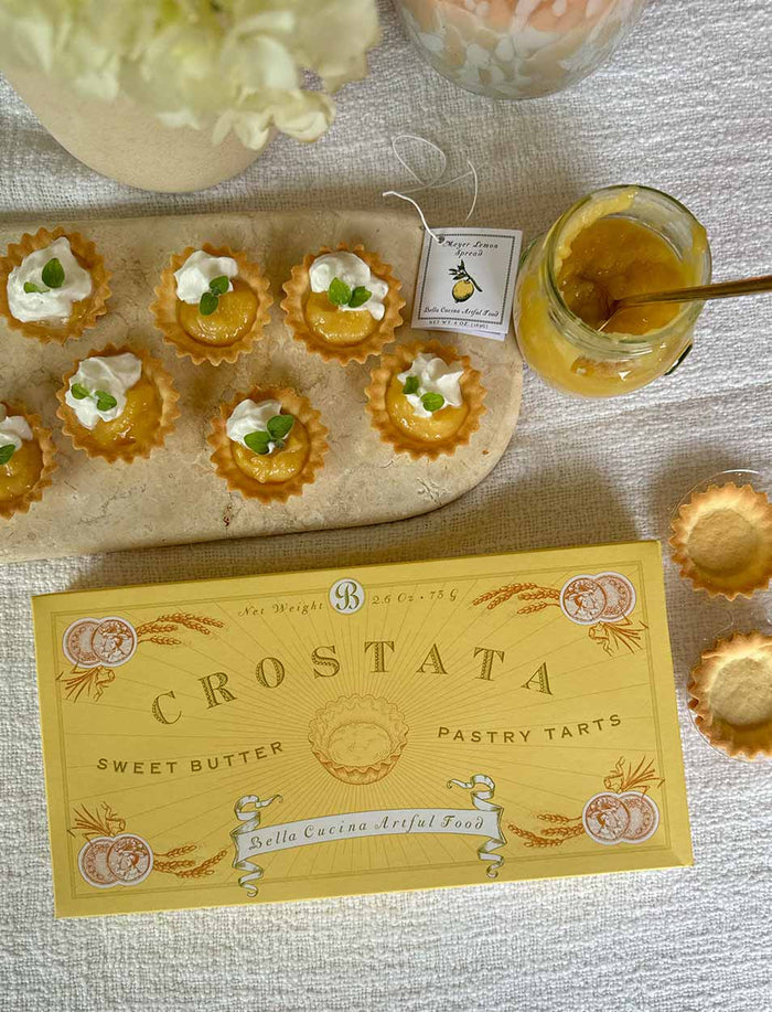 A yellow Crostata pastry tart box sits on a table beside mini tarts filled with cream and herbs, an open jar of Bella Cucina Meyer Lemon Spread with a spoon, and empty tart shells on a light textured surface.