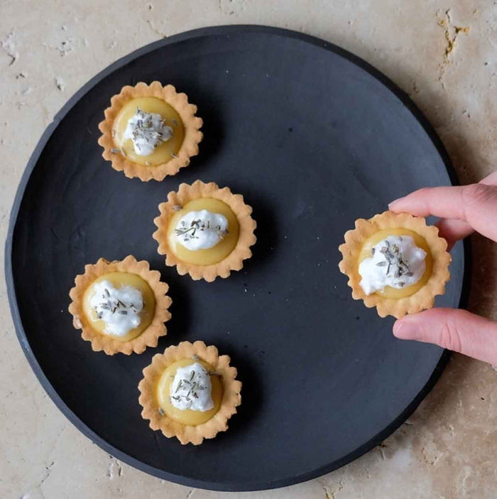 Five small tartlets topped with cream, herbs, and Bella Cucina Meyer Lemon Spread are arranged on a round black plate. A hand reaches for one tartlet from the plate, which rests on a light textured surface.