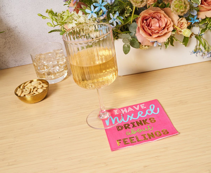 A glass of white wine rests on a Sophistiplate 'Mixed Drinks About Feelings' cocktail napkin (26pk), beside a bowl of nuts, another drink, and a floral bouquet on a wooden table.