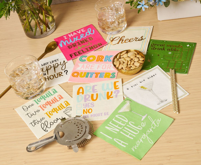 A tabletop with two whiskey glasses, a cocktail strainer, a bowl of nuts, and Sophistiplate's 'Mixed Drinks About Feelings' Cocktail Napkins (26pk) featuring humorous drink quotes. A gold spoon, flowers, and festive gold foil napkins complete the lively scene.