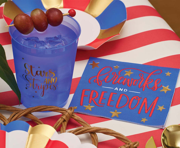 A blue cup with metallic gold accents reading “Stars and Stripes” sits on a red-and-white striped tablecloth beside Sophistiplate’s Modern Patriotic Cocktail Napkins Fireworks/20pk; the cup holds ice and grape skewers amid festive decor.