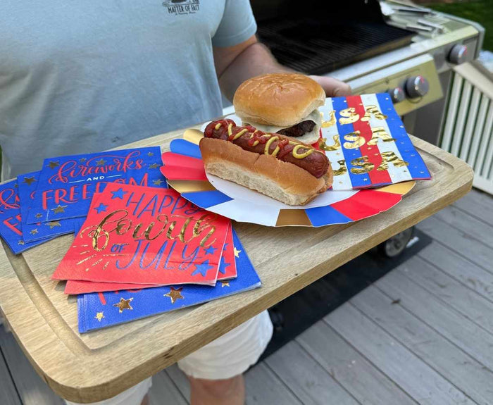 On a deck near a grill, someone holds a tray with a cheeseburger, hot dog with ketchup and mustard, and Sophistiplate Modern Patriotic Guest Towels/20pk featuring metallic gold accents.