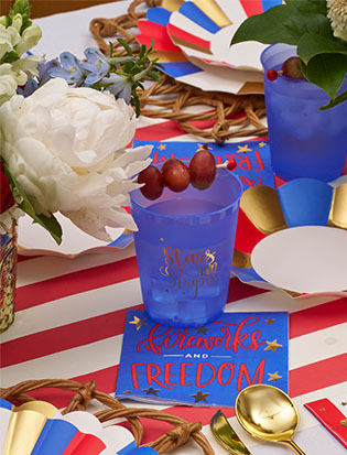 A festive table is set with a red and white striped tablecloth, blue cups with gold accents, plates, grapes, white flowers, and Sophistiplate's Modern Patriotic Cocktail Napkins Fireworks/20pk for a patriotic touch.