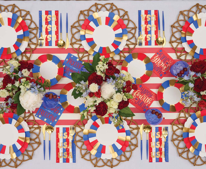 A festive table features red, white, blue, and gold accents with striped placemats, floral centerpieces, gold utensils, and Sophistiplate’s Modern Patriotic Guest Towels. Plates have multicolor borders and toast to USA and Happy Fourth.