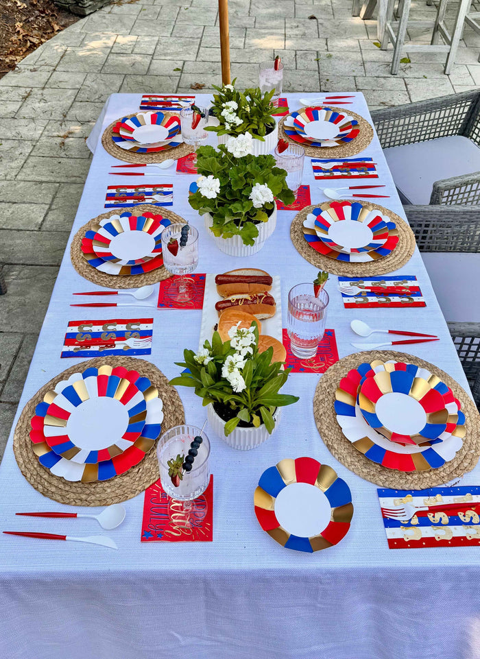 A rectangular outdoor table set for eight features Sophistiplate Modern Patriotic Dinner Plates (8pk), woven placemats, red napkins, white potted flowers, hot dogs, and drinks for a festive scene with red, white, blue, and gold accents.