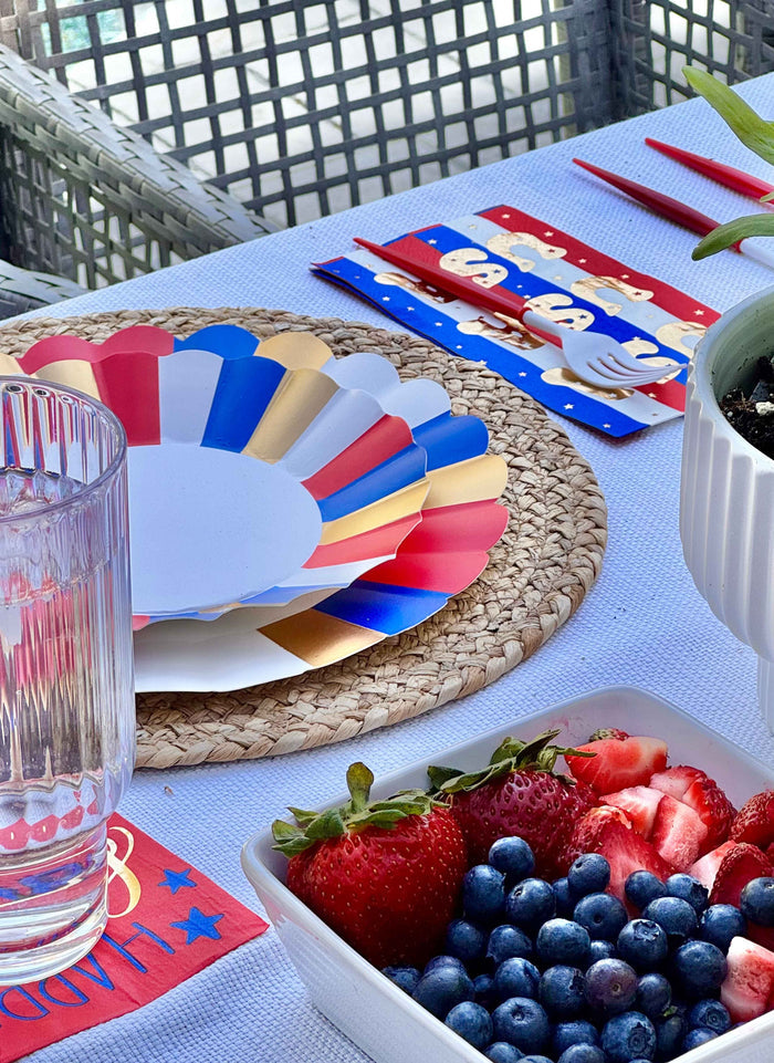 A festive table features Sophistiplate's Modern Patriotic Dinner Plates/8pk with scalloped red, white, blue, and gold edges, a glass of water, cutlery on a patriotic napkin, and a bowl of strawberries and blueberries on a woven placemat.