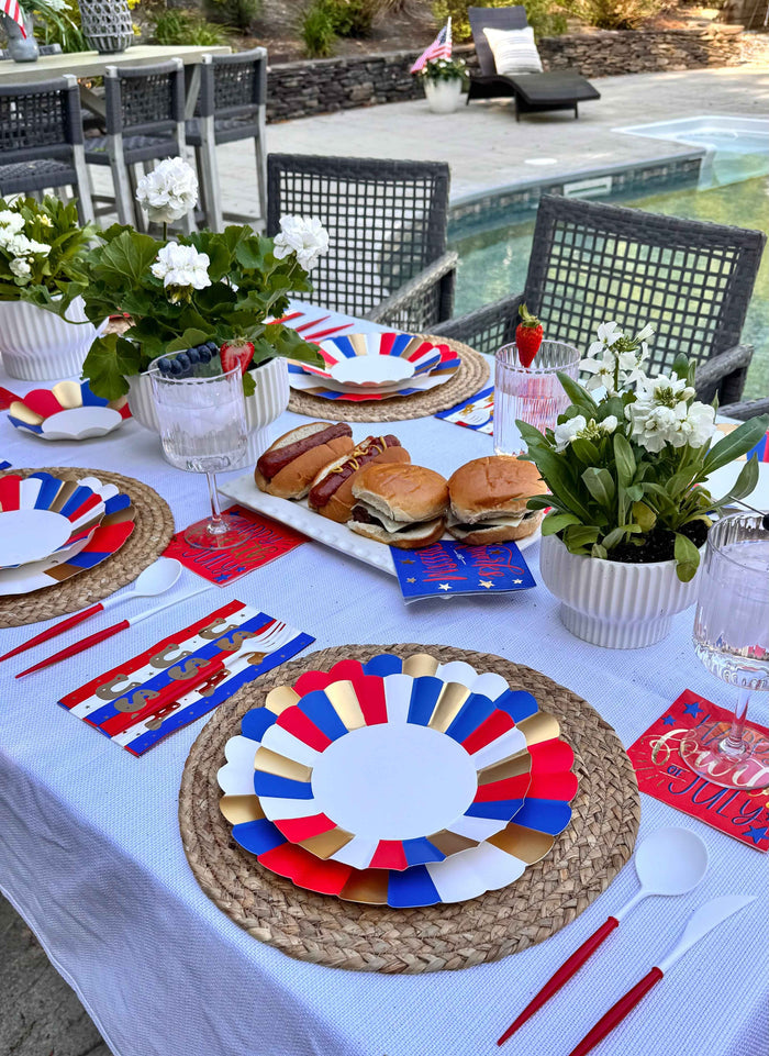 A festive outdoor poolside table features Sophistiplate Modern Patriotic Dinner Plates (8pk), burgers, drinks, napkins, and white floral centerpieces for a vibrant red, white, and blue holiday vibe.
