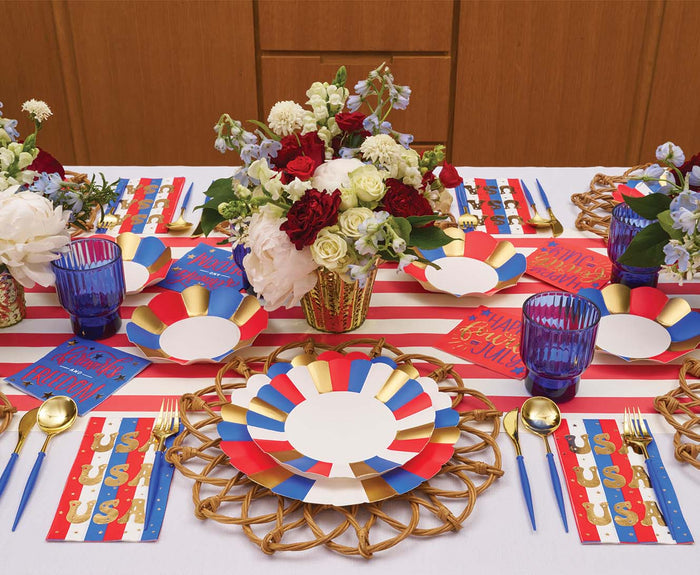 A festive table setting with Sophistiplate Modern Patriotic Salad Plates (8pk), blue glasses, gold cutlery, striped placemats, USA-themed napkins, disposable paper plates, and floral centerpieces with red and white flowers.