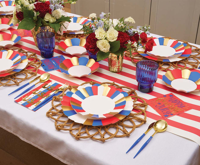 A festive table is set with Sophistiplate Modern Patriotic Salad Plates, gold accents, woven placemats, blue glasses, colorful cards, and fresh flowers in vases atop a red and white striped runner.