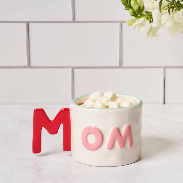 The Mom Mug + Cocoa Gift Set by Sophistiplate LLC features a white mug with pink and red letters, filled with marshmallows—an ideal gift for mom or as part of a hot cocoa set. Displays beautifully on a counter with flowers.