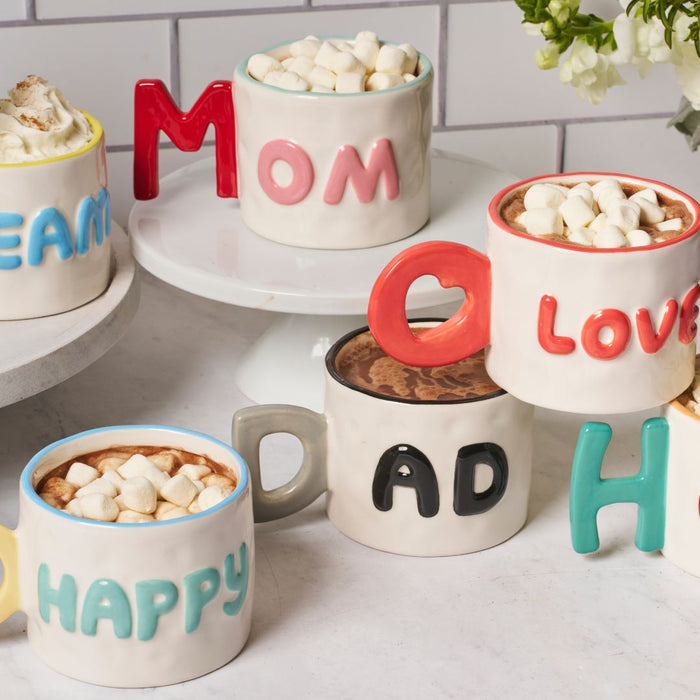 Five colorful mugs from the Sophistiplate LLC Dad Mug + Hot Cocoa Gift Set, made with Bang Candy Hot Cocoa Mix and topped with marshmallows, are arranged on a white surface. Each mug spells MOM, LOVE, HAPPY, DREAM, or DAD.