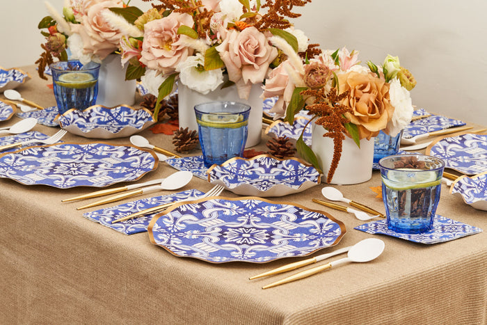The Moroccan Table Setting by Sophistiplate LLC features blue and white patterned heavy-duty paper plates, gold cutlery, blue glasses, and vases with pink and orange flowers on a beige tablecloth.
