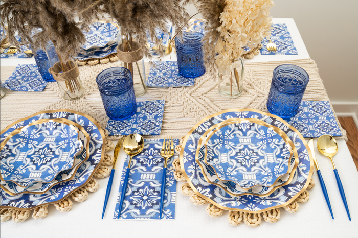 The Moroccan Table Setting by Sophistiplate LLC features blue and white patterned heavy-duty paper plates and napkins, gold utensils, blue glasses, woven placemats, beige macrame decor, and dried floral centerpieces.