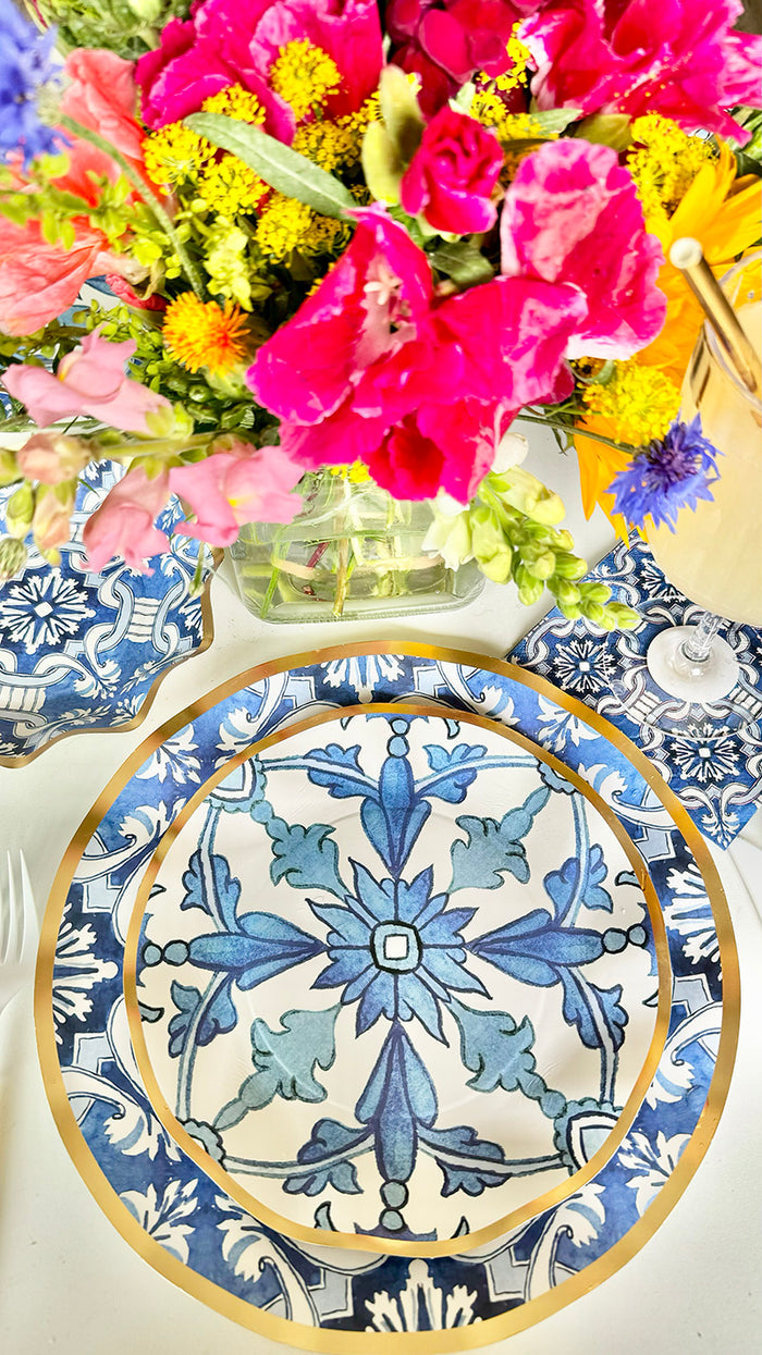 A vibrant floral arrangement sits above a Moroccan Table Setting by Sophistiplate LLC, featuring a decorative blue and white plate with gold trim, a coordinating napkin, and part of a wine glass in view.