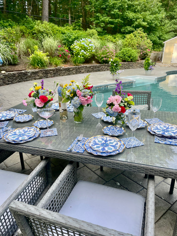 The Moroccan Table Setting by Sophistiplate LLC features blue and white heavy-duty paper plates, napkins, glassware, cutlery, and colorful floral accents—a perfect poolside look with lush plants and a stone wall in the background.