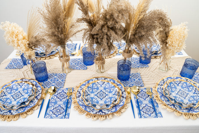 The Moroccan Table Setting by Sophistiplate LLC features blue and white disposable plates, gold cutlery, blue glasses, beige placemats, and a centerpiece with dried pampas grass and flowers in small vases for an elegant bohemian look.
