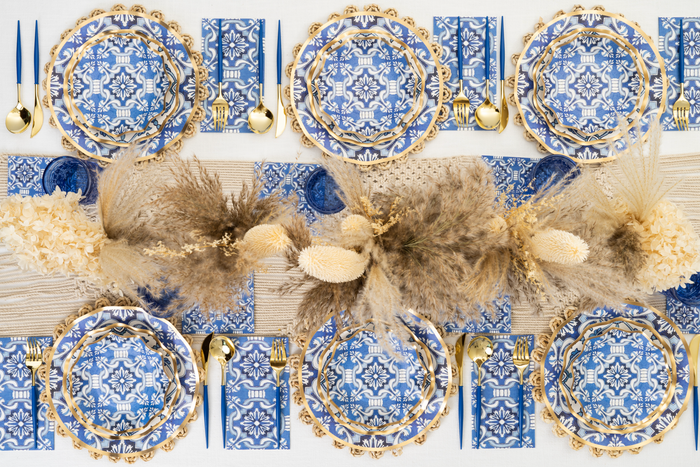 A table set for a meal with blue and white patterned plates, gold cutlery, blue glasses, and blue patterned napkins. A centerpiece of dried beige grasses and flowers runs down the middle of the table.