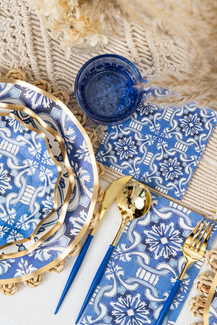 The Moroccan Table Setting by Sophistiplate LLC features blue and white patterned plates, napkins, gold and blue cutlery, a blue glass, and a woven placemat—perfect for a stylish party-in-a-box.