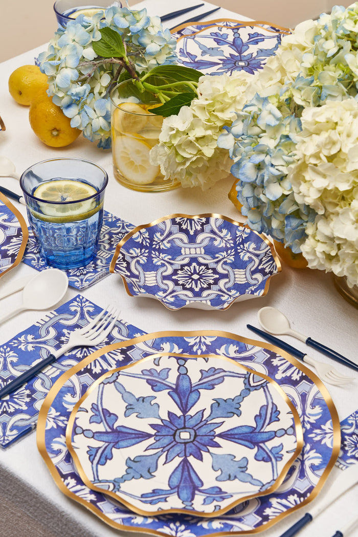 Create a summer table with Sophistiplate’s Moroccan Nights Wavy Paper Salad Plates, gold accents, blue glasses of lemon water, navy utensils, hydrangea vases, and fresh lemons on a white tablecloth.