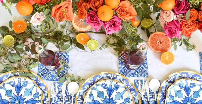 The Moroccan Table Setting by Sophistiplate LLC features blue and white disposable plates, floral napkins, cutlery, soda bottles, and a centerpiece with vibrant flowers and citrus on a white tablecloth.