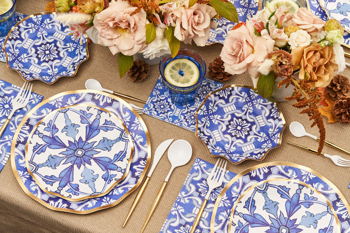 The Moroccan Table Setting by Sophistiplate LLC features blue and white heavy-duty paper plates, gold flatware, white-handled utensils, floral displays with pink and peach roses, pine cones, lemon drinks on blue coasters, and a burlap tablecloth.