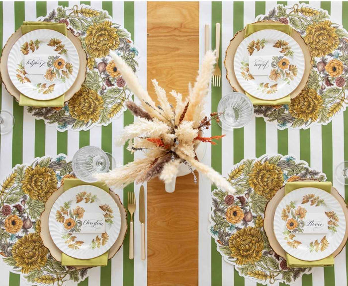 A top-down view of a wooden dining table set for four with green and white striped placemats, floral plates, gold cutlery, clear glasses, and the Hester & Cook Moss Classic Stripe Runner (20x25") as a base. Name cards sit on each plate; dried flowers form the centerpiece.