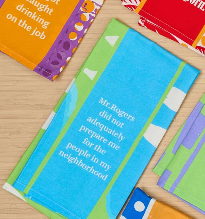 The Mr Rogers Dish Towel by Drinks On Me features the witty quote "Mr. Rogers did not adequately prepare me for the people in my neighborhood," making a humorous kitchen gift, shown with other patterned napkins on a light wood surface.