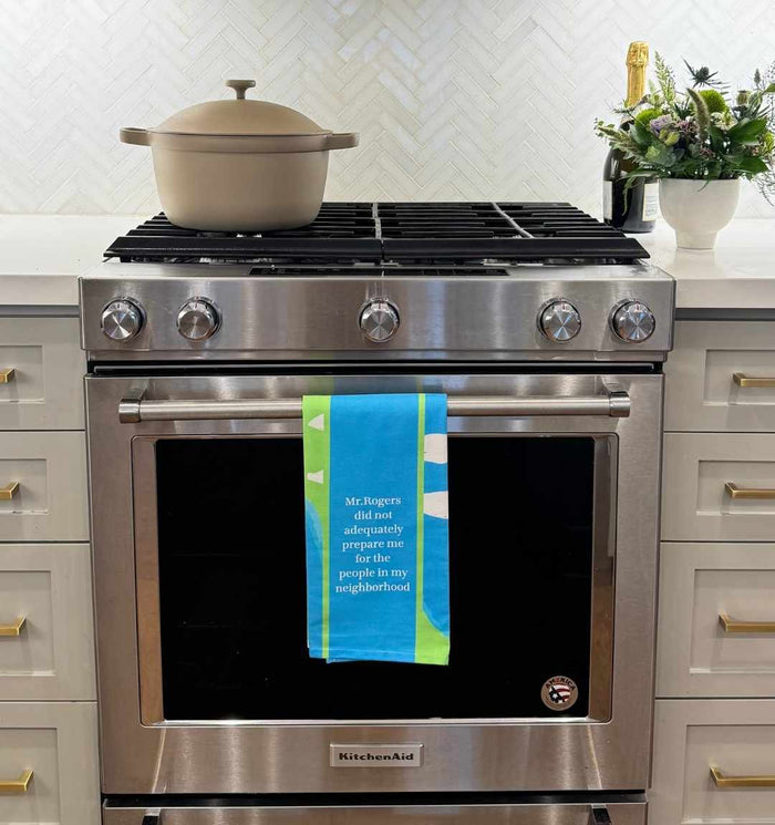 A Mr Rogers Dish Towel by Drinks On Me hangs on a stainless steel oven, featuring the quote, "Mr. Rogers did not adequately prepare me for the people in my neighborhood." A beige pot sits on the stove.