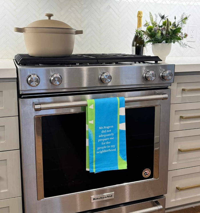 A Drinks On Me Mr Rogers Dish Towel hangs on a stainless steel KitchenAid oven, reading, "Mr. Rogers did not adequately prepare me for the people in my neighborhood"—a perfect humorous kitchen gift.