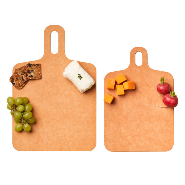 Two Maple Natural Wood Fiber Composite Boards with handles are spaced apart on a white background. The left board holds grapes, crackers, and cheese; the right has cubed orange cheese and radishes. Both are heat-resistant.