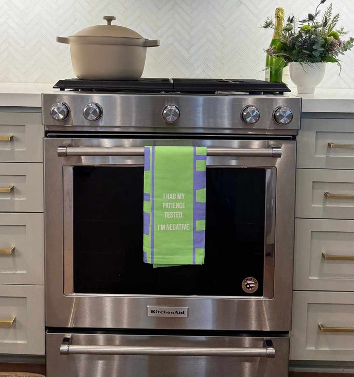 A stainless steel KitchenAid oven features a green and blue Drinks On Me Negative Dish Towel hanging on the handle that reads, "I had my patience tested. I’m negative." A pot and a vase with flowers sit on the white countertop above.
