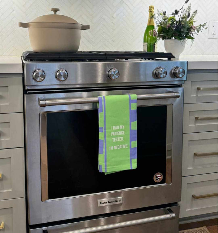 A stainless steel oven displays the Drinks On Me Negative Dish Towel in green and blue, reading "I had my patience tested. I'm negative." A beige pot is on the stove, with flowers and a bottle on the counter.