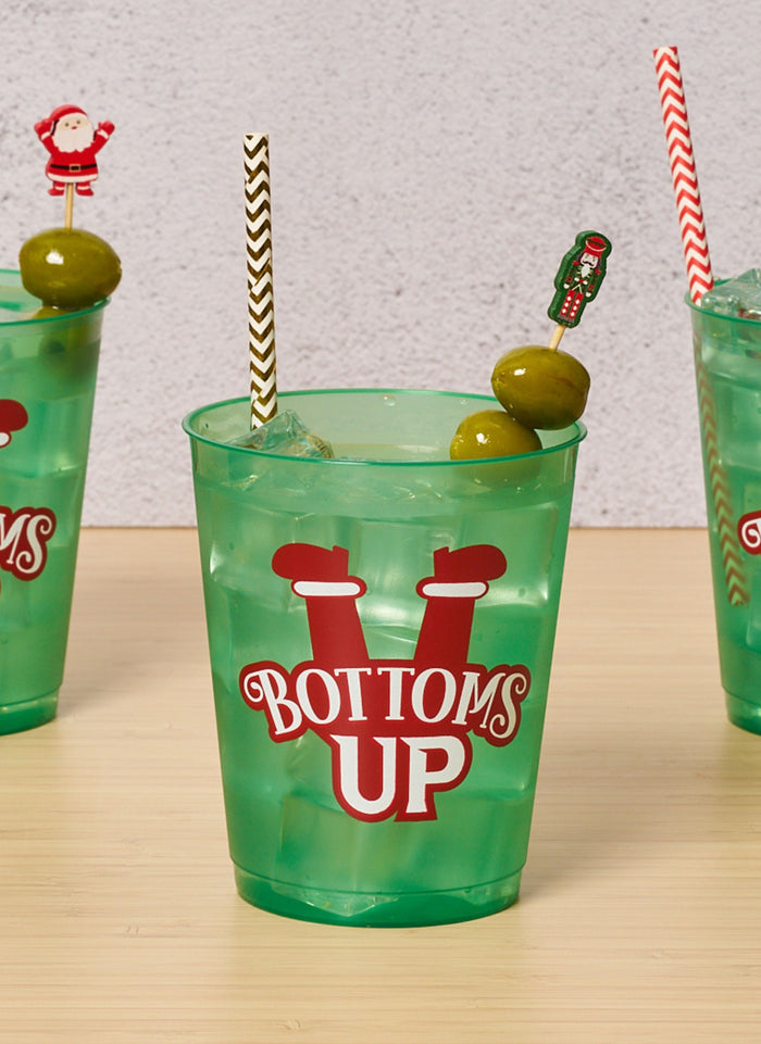 A green plastic cup labeled Bottoms Up holds an iced drink, garnished with two green olives on Simply Serv'd Nutcracker Christmas Large Cocktail Picks, plus two patterned straws. Two similar cups appear partially in the background.