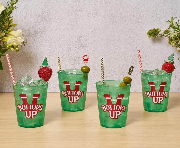 Four green cups with BOTTOMS UP text, each filled with drinks and topped with a festive Simply Serv'd Nutcracker Christmas Large Cocktail Pick, sit on a wooden surface surrounded by flowers and greenery.