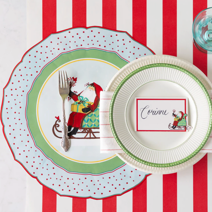A festive table features a red and white striped placemat, Santa plates, cutlery, napkin, water glass, and a Hester & Cook Old St. Nick Place Card from the 12ct set, adding charm to your Christmas decor.