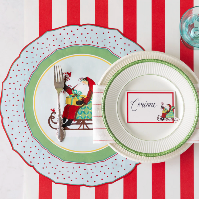 A festive place setting featuring Hester & Cook’s Old St. Nick Placemats (12ct), a Santa-themed plate, cutlery, a glass of water, and a name card—perfect for cheerful holiday table décor.
