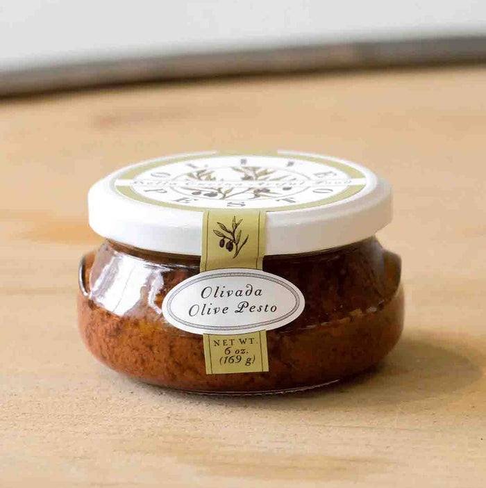 A small glass jar of Bella Cucina Olivada Olive Pesto with a white lid and cream label sits on a light wooden surface, filled with dark, chunky Kalamata olive Mediterranean pesto spread.